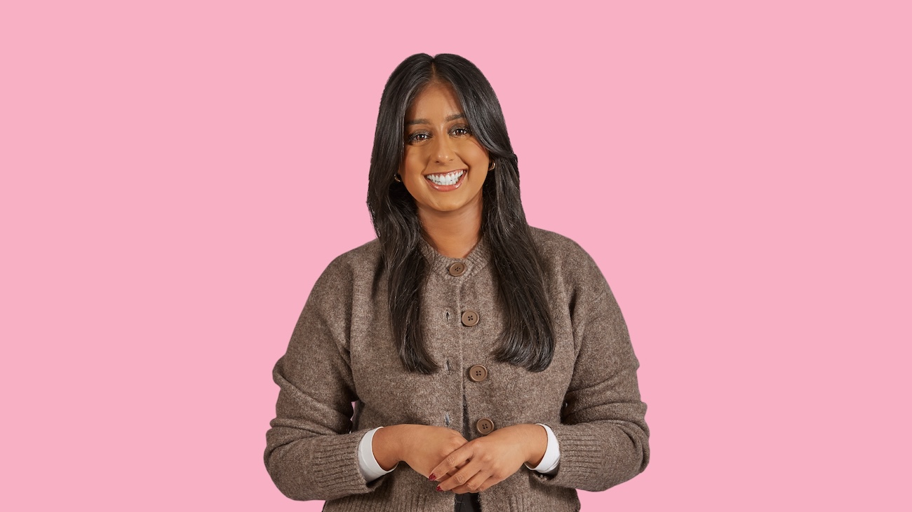 Woman with long dark hair, wearing a brown sweater, smiling on a pink background.