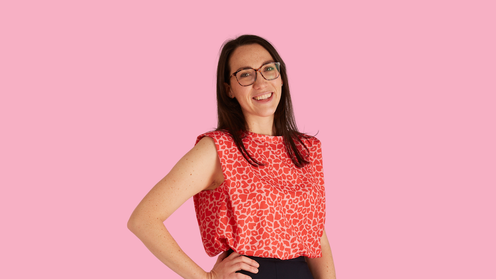 Woman with glasses smiling, wearing a red patterned top, pink background.