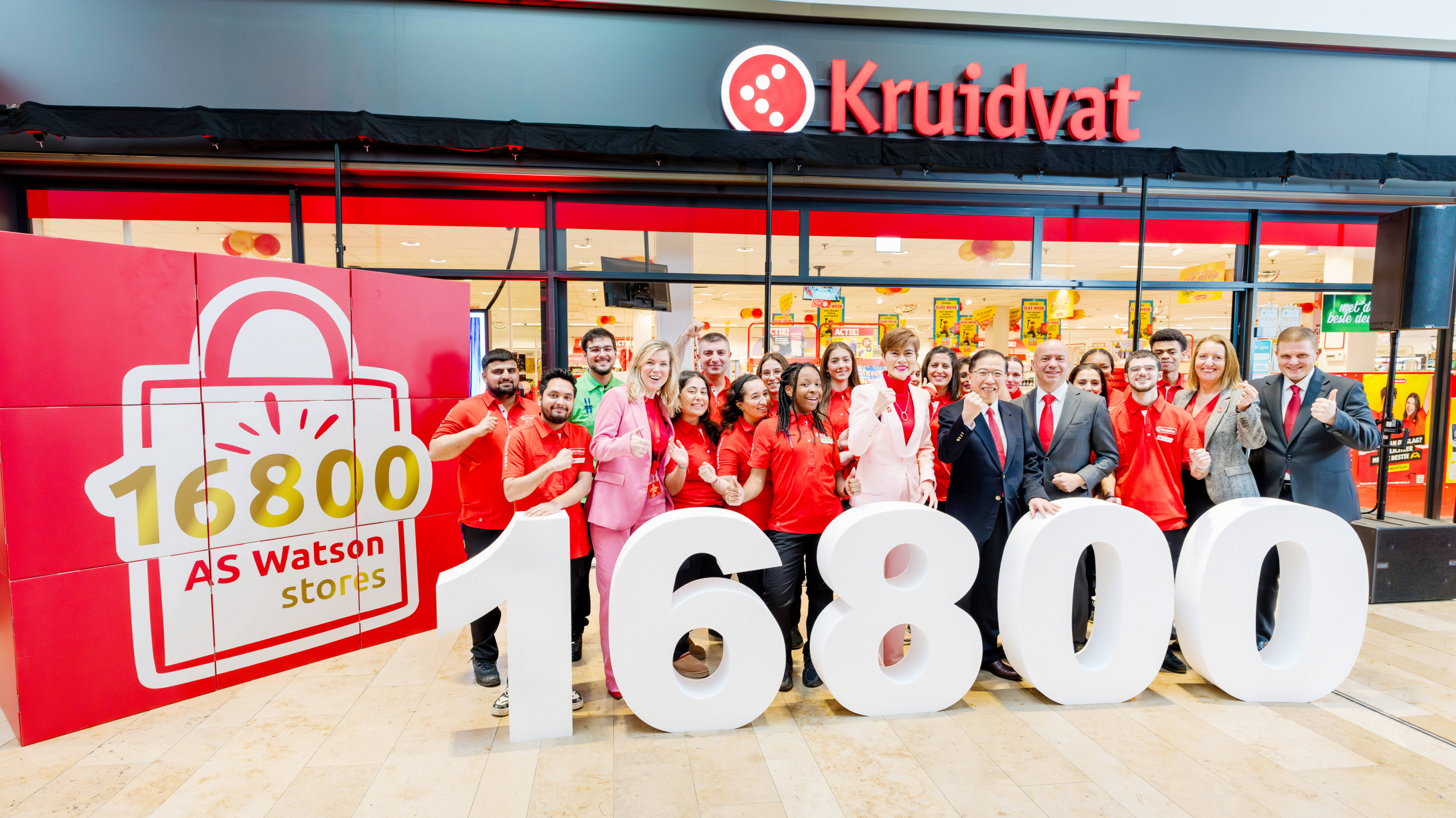 Group of people celebrating in front of a Kruidvat store, with a sign that says "16800 AS Watson stores".