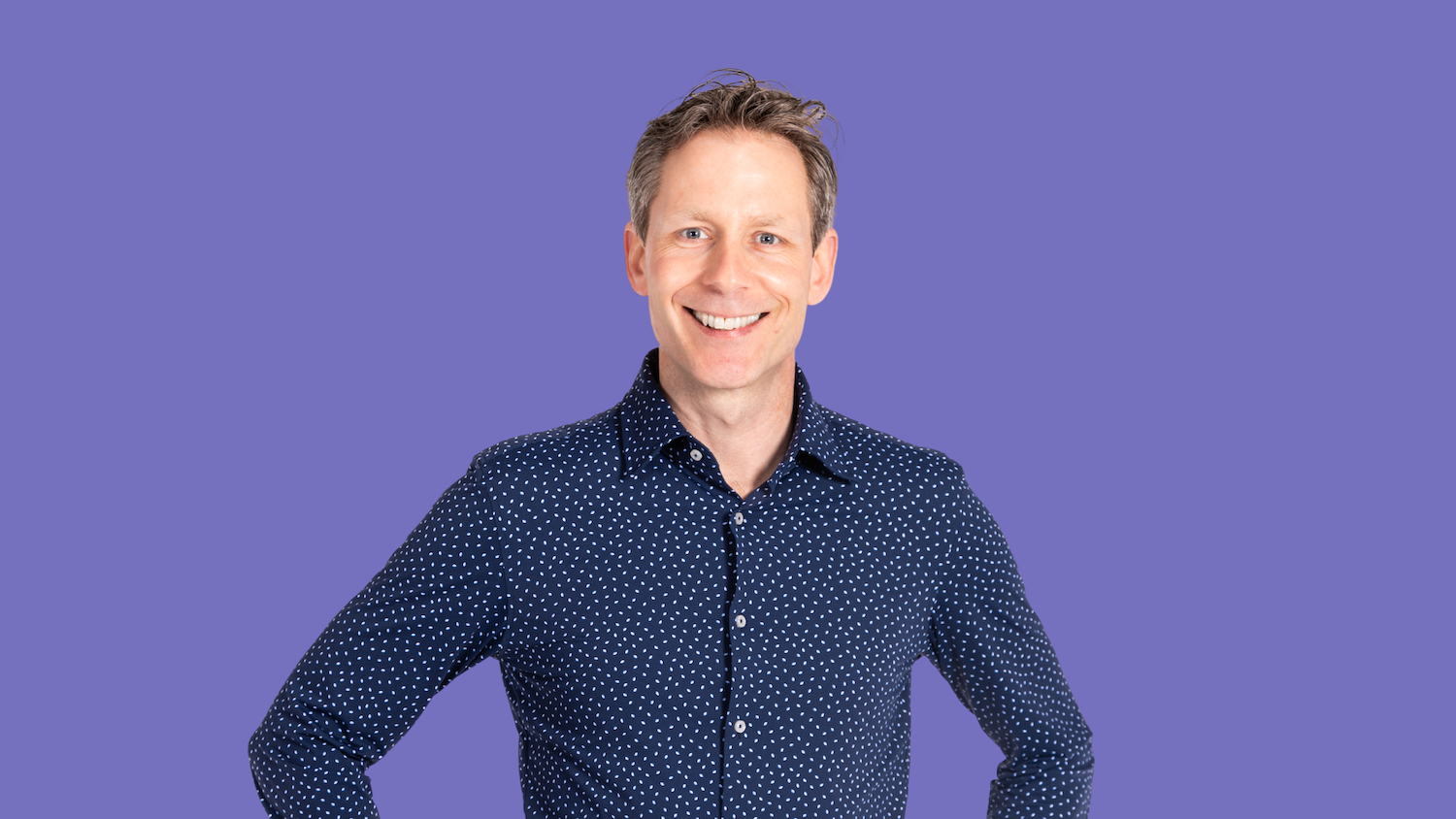 Man with a blue patterned shirt smiling on a purple background.