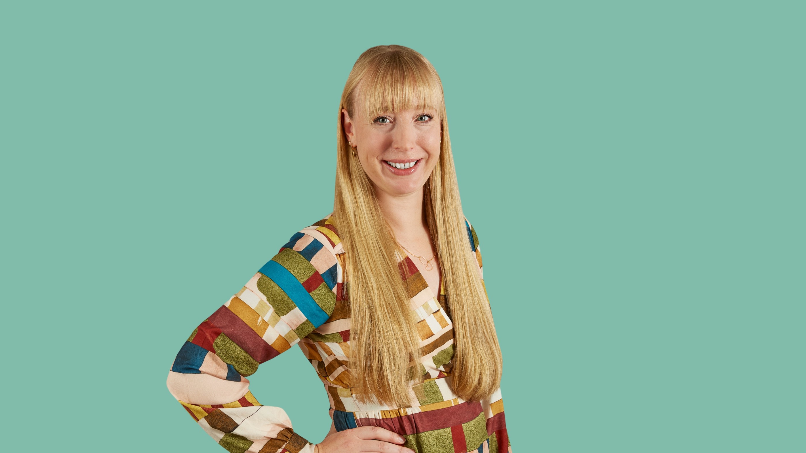 Woman with long blonde hair smiling, wearing a colorful dress, and posing on a teal background.