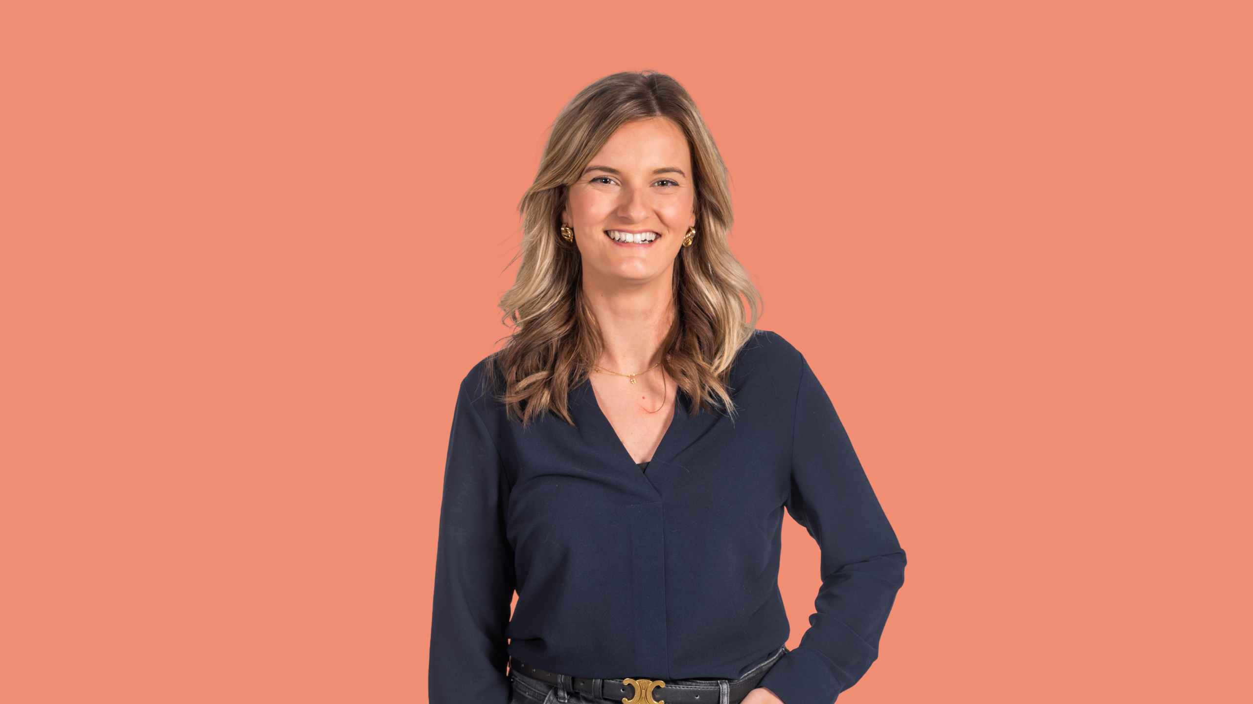 Woman with blonde hair wearing a navy blue top, smiling, and posing against a peach background.