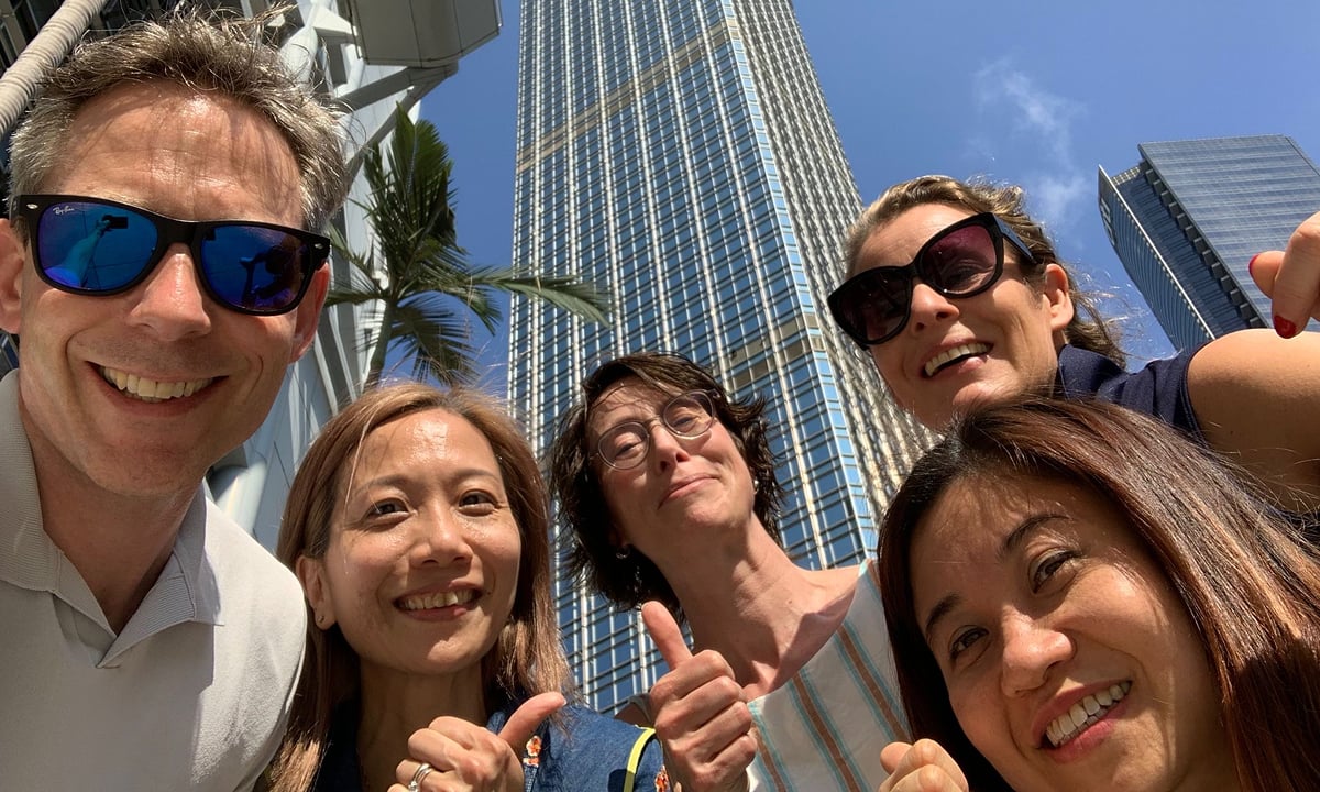 Group of people smiling, thumbs up, tall buildings in background.