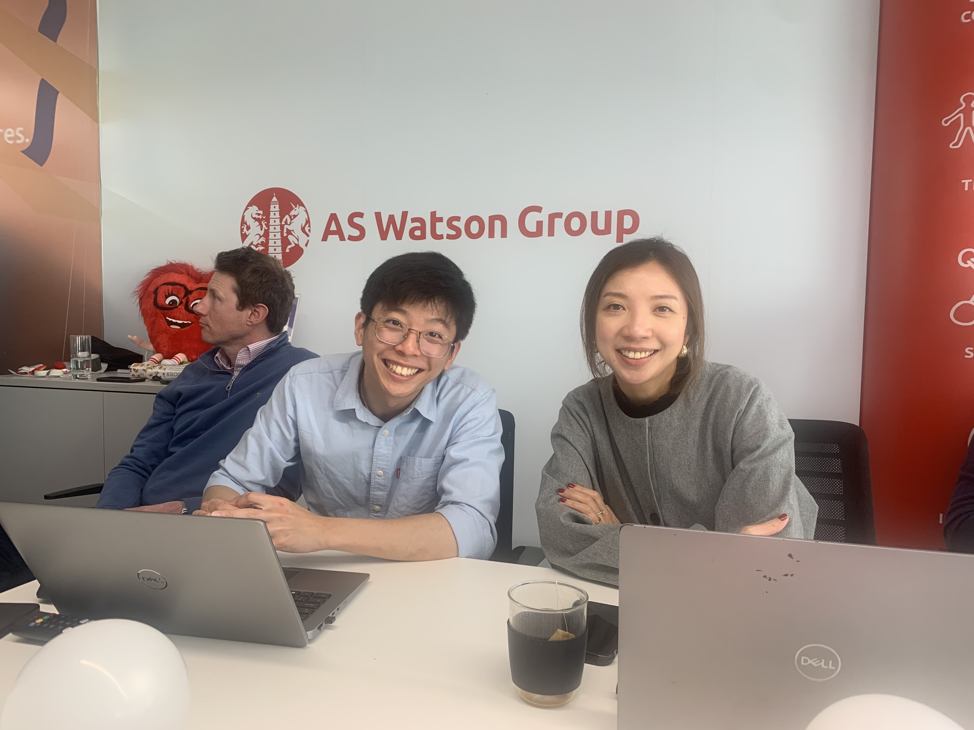 Three people at a table with laptops, smiling. AS Watson Group logo.