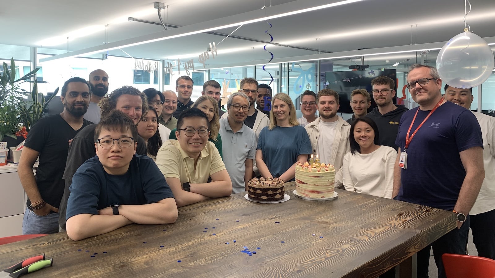 Group of people around a table with two cakes, celebrating a birthday.