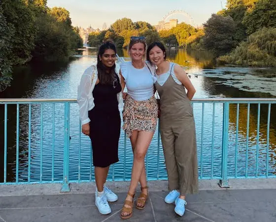 Three women posing on a bridge, water and trees in the background.