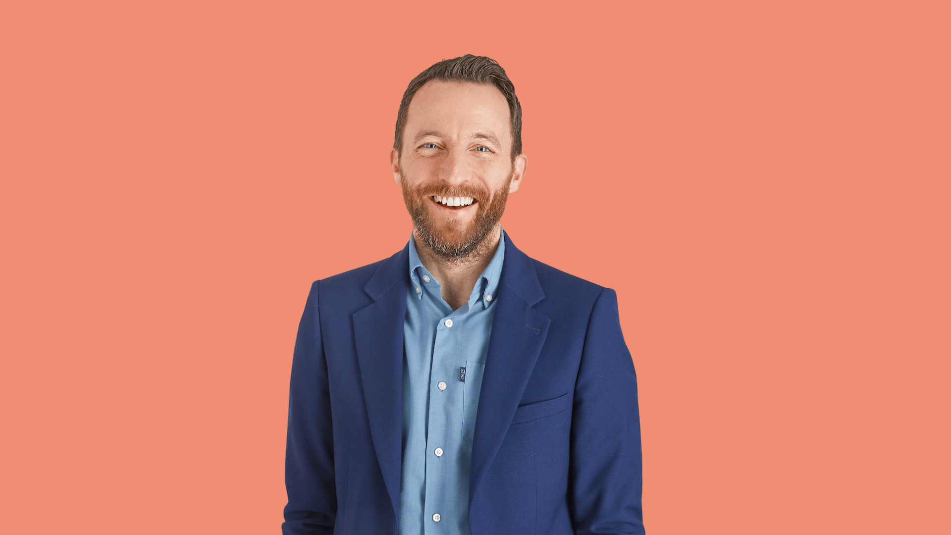 Smiling man in a blue suit and light blue shirt, coral background.