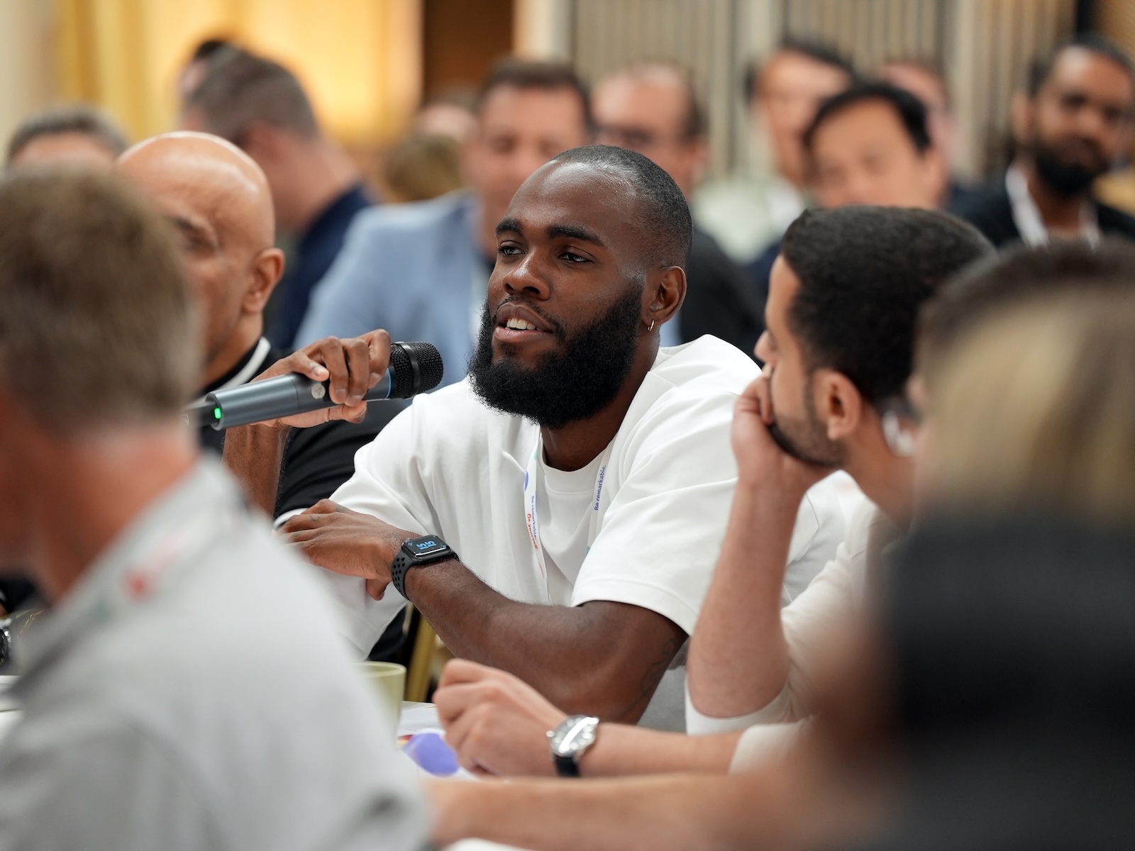 Man with microphone, speaking at a conference, surrounded by other attendees.