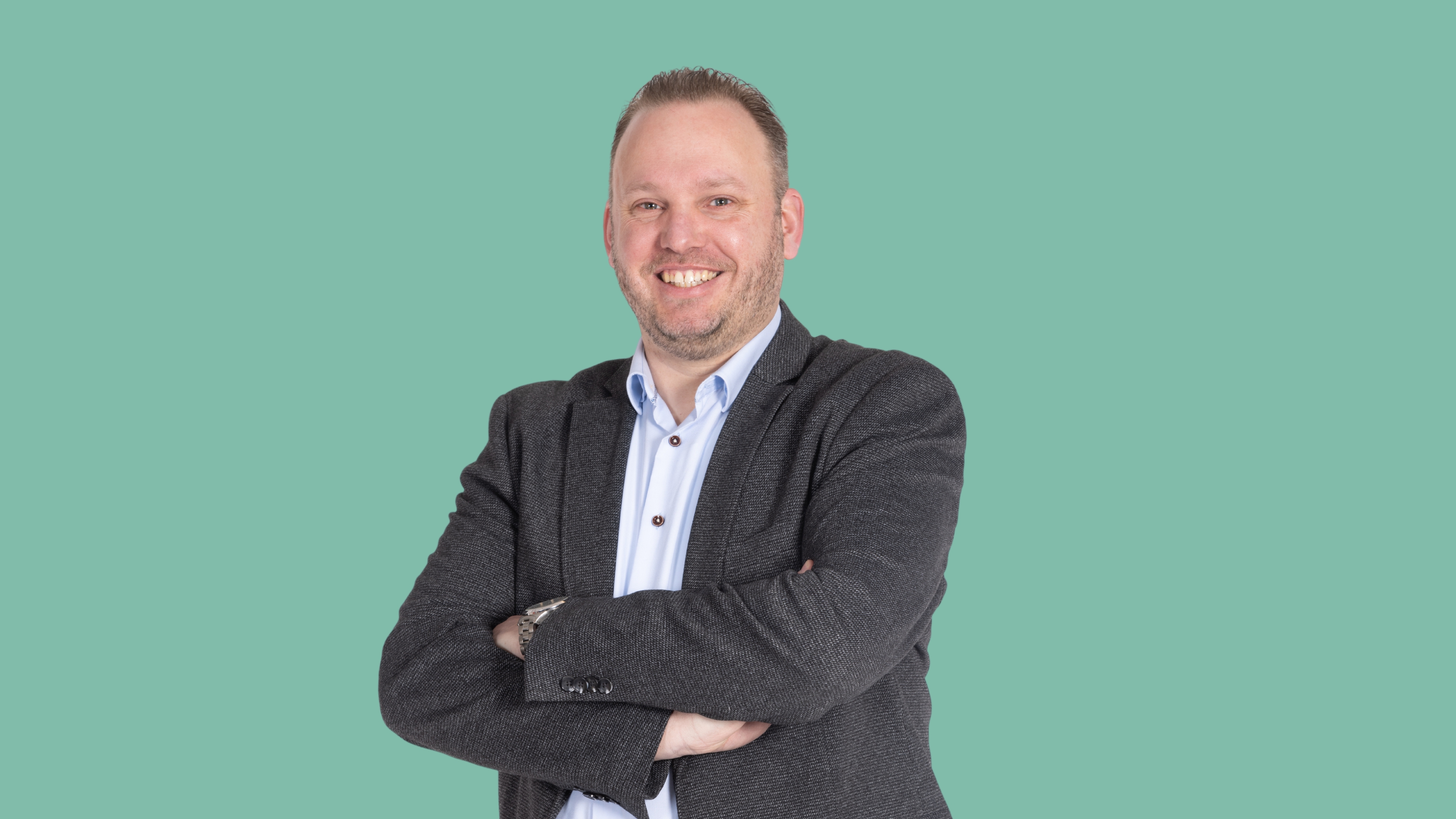 Man in blazer with arms crossed, smiling, on a teal background.