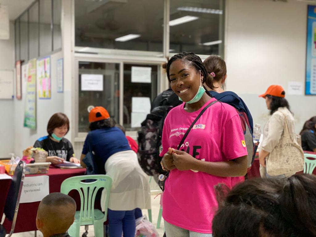 Woman in pink shirt, smiling, wearing a face mask and braids. Other people in the background.
